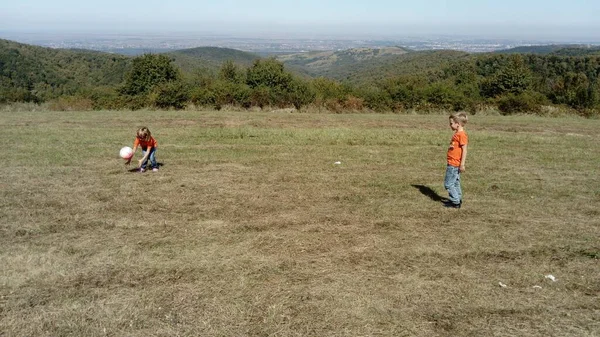 Un garçon et une fille aux cheveux blonds, vêtus de t-shirts orange et de jeans classiques, jouent au ballon dans une prairie de montagne. Début automne chaud. Les enfants jouent. En arrière-plan l'horizon et la végétation — Photo