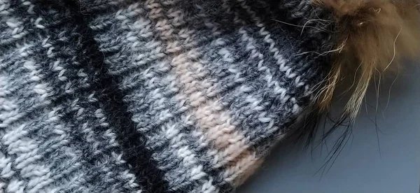 Strickmütze mit einem Kürbis in Großaufnahme auf grauem Hintergrund. Winterbekleidungswerbung. Wintersaison. Weißgrau, Beige, Braun und Schwarztöne. Große Schleifen. der bohnenbraune Kürbis aus natürlichem Fell — Stockfoto