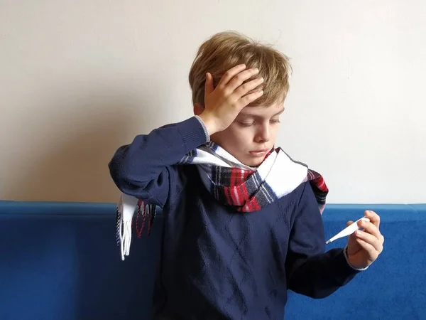 El chico se enfermó, le duele la cabeza. La enfermedad viral está progresando. El cuello de los niños está envuelto en una bufanda caliente. El niño mira abatido al termómetro, sostiene su mano sobre su frente y sufre — Foto de Stock