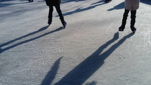 Los niños viajan en un parque de la ciudad en una pista de hielo. Patinador de pies mientras patina sobre hielo. El bajo sol de invierno ilumina débilmente el hielo. Formas oscuras y largas sombras en la superficie. Movimientos deportivos — Foto de Stock