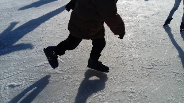 Kinder reiten im Stadtpark auf einer Eisbahn. Füße Skater beim Schlittschuhlaufen auf Eis. die tief stehende Wintersonne beleuchtet das Eis schwach. dunkle Formen und lange Schatten auf der Oberfläche. Sportbewegung — Stockfoto