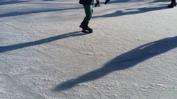 Los niños viajan en un parque de la ciudad en una pista de hielo. Patinador de pies mientras patina sobre hielo. El bajo sol de invierno ilumina débilmente el hielo. Formas oscuras y largas sombras en la superficie. Movimientos deportivos — Foto de Stock