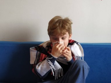 Çocuk hastalandı, boğazı ağrıyor ve vücut ısısı yükseliyor. Çocukların boynu sıcak bir eşarba sarılmış. Çocuk burnunu siliyor, gözlerini şişiriyor. Beyaz saçlı, sarı saçlı çocuk..