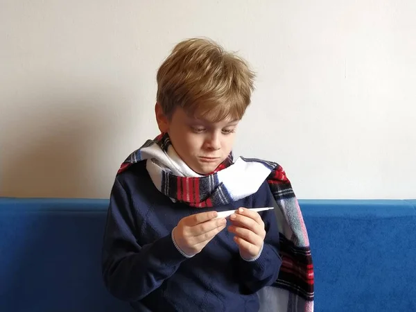 The boy got sick, his throat hurts and his body temperature rises. The childs neck is wrapped in a warm scarf. The kid looks dejectedly at the thermometer and is surprised — ストック写真