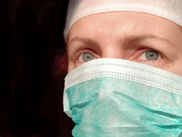 Woman doctor or nurse in green protective mask. Red hair, black background. A worried look into the distance. Middle-aged or elderly woman with the lower half of her face hidden under a mask.