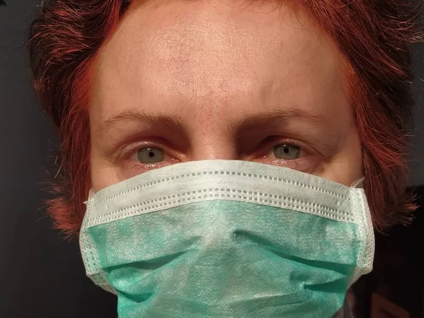 Redhead woman with sick tired eyes in a protective surgical mask of green color. Gray eyes and red dyed hair. Dark background. Protective measures against viral and bacterial diseases. Anxious look.