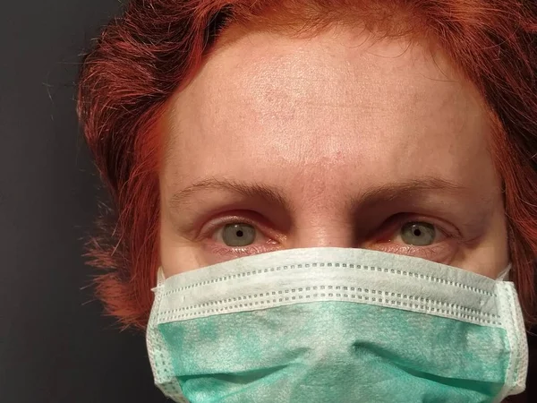 Redhead woman with sick tired eyes in a protective surgical mask of green color. Gray eyes and red dyed hair. Dark background. Protective measures against viral and bacterial diseases. Anxious look.