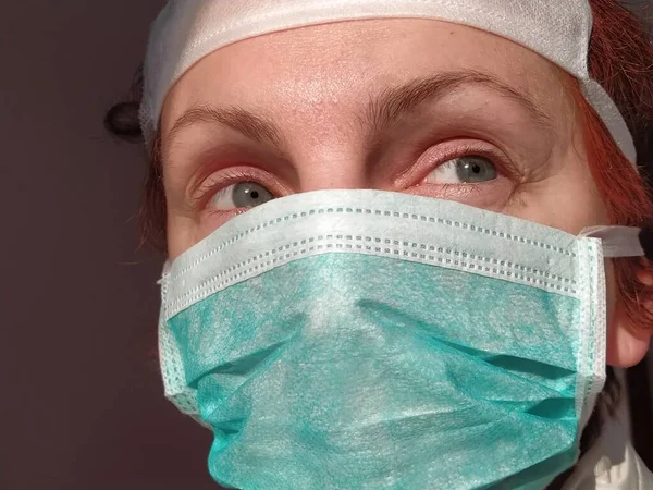 Redhead woman with sick tired eyes in a protective surgical mask of green color. Gray eyes and red hair. Dark background. An alarming look to the side and up. Sly squint or grimace on the face