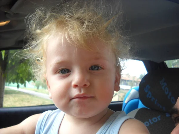 One year old baby in a car. A boy of a white race with blond fluffy, shaggy hair. Cute kid portrays the father of the car. Summer clothes. Blue T-shirt. The tradition of not cutting hair for toddler. — Stock Image One Year Old Baby Car Boy White Race Blond Fluffy Royalty Free Stock Photos