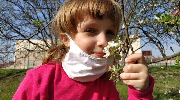 Cute white girl of 6 years old takes off a white surgical protective mask and sniffs beautiful fragrant flowers on an apple tree. The concept of recovery from a serious illness.