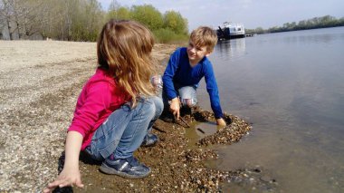 Erkek ve kız kardeş nehir kenarında oynuyorlar. Bir erkek ve bir kız kot pantolon ve uzun kollu kıyafetler giyiyorlar. Yazın başında. Gülümse ve su kenarında oyna. Pembe bluz ve mavi uzun kollu. Sırbistan