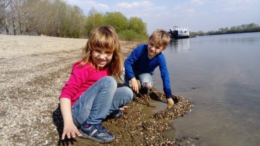 Erkek ve kız kardeş nehir kenarında oynuyorlar. Bir erkek ve bir kız kot pantolon ve uzun kollu kıyafetler giyiyorlar. Yazın başında. Gülümse ve su kenarında oyna. Pembe bluz ve mavi uzun kollu. Sırbistan