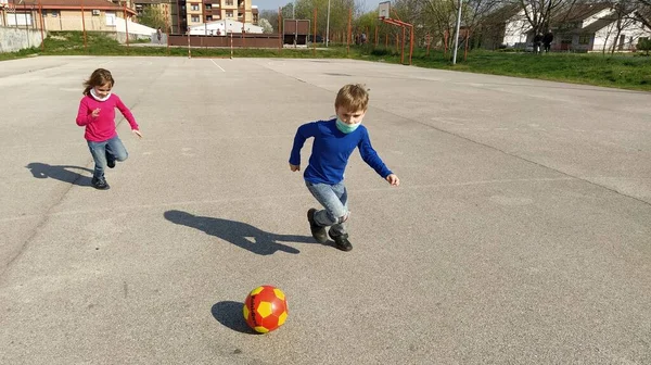 女子と男子はアスファルト競技場でサッカーをする 子供たちは空気感染症を防ぐために保護外科マスクを着用する ウイルスのパンデミックの間の家族スポーツ 子供たちはジーンズを着ています 晴れた日 — ストック写真