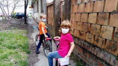 Children in white protective surgical masks go in for cycling. Pause in cycling. Girl 6 years old in a pink T-shirt and a blond boy 7 years old in orange. Kids on the street near the old brick fence. clipart