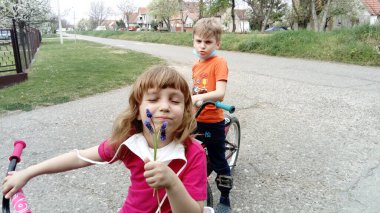 Belgrade, Serbia, April 13, 2020: children in protective surgical masks ride bicycles. The kids took off their masks. The girl sniffs a purple flower and smiles, and the boy frowns an eyebrow and says something to her.