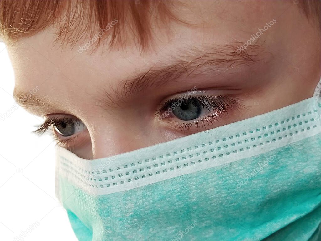 Lovely white boy in a protective surgical mask of green color. Close-up of the head and face. Glance lowered down and to the side. Gray eyes and long eyelashes. White background. Prevention of airborne diseases.