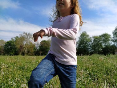 Yeşil çimlerle tarlada koşan bir kız. Çocuk beyaz bir bluz ve kot pantolon giyiyor. Yaz akşamı. Çocuk eğlencesi. Uzun sarı saçlı kız..