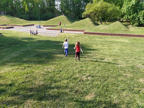 Сремска-Митровица, Сербия - 24 апреля 2020 года - Дети бегают по парку. Мальчик в белой футболке и синих брюках. Девушка в красной майке и брюках. Зеленая лужайка или поле. Люди гуляют в городском парке — стоковое фото