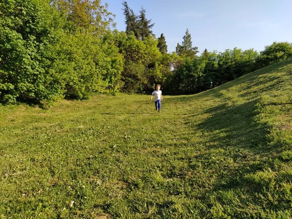 V parku pobíhá dítě. Chlapec v bílém tričku a modrých kalhotách. Zelený trávník nebo pole. Sekaný trávník. Procházka a hrát si v lese. Aktivní sportovní aktivity. — Stock fotografie