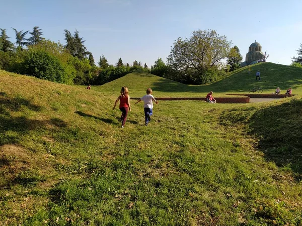 Sremska Mitrovica, Srbsko - 24. dubna 2020 - V parku běží děti. Chlapec a dívka hrají venkovní hry. Zadní pohled. Zelený trávník nebo pole. Lidé chodí do městského parku — Stock fotografie