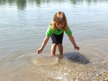 Güneşli bir yaz gününde güzel bir nehirde su sıçratan sevimli kıvırcık kız. Çocuk yeşil tişört ve siyah şort giyiyor.