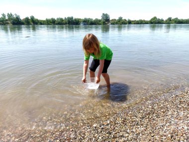 Güneşli bir yaz gününde güzel bir nehirde su sıçratan sevimli kıvırcık kız. Çocuk yeşil bir tişört ve siyah bir şort giydi. Sırbistan, Sava Nehri ve Sremska Mitrovica.
