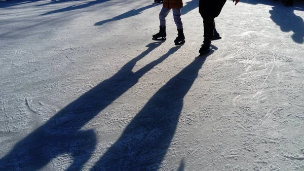 子供たちはアイスリンクの都市公園に乗っています アイススケートをしながら足のスケーター 低い冬の太陽は氷を弱く照らす 表面の暗い形と長い影 — ストック写真