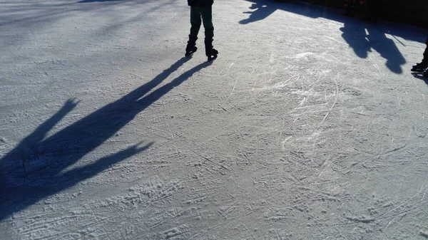 子供たちはアイスリンクの都市公園に乗っています アイススケートをしながら足のスケーター 低い冬の太陽は氷を弱く照らす 表面の暗い形と長い影 — ストック写真