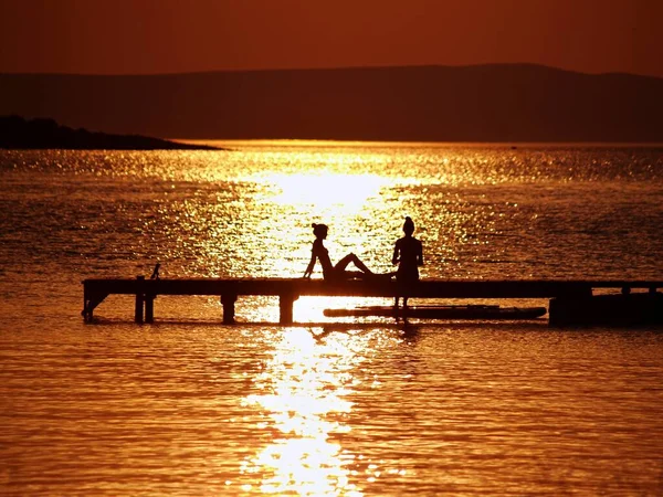 İki genç, sıska, formda kızın siluetleri, güneşin altında bir kıyıda dinleniyor, altın saatinde su üzerinde parıldıyor, terk edilmiş Kampor Koyu 'nda, ada Haham' da.