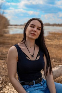 Güzel bir kız nehir kıyısında bir kütüğün üzerinde oturuyor kot pantolon ve siyah tişörtle.