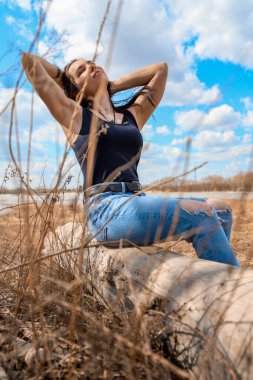 Güzel bir kız nehir kıyısında bir kütüğün üzerinde oturuyor kot pantolon ve siyah tişörtle.