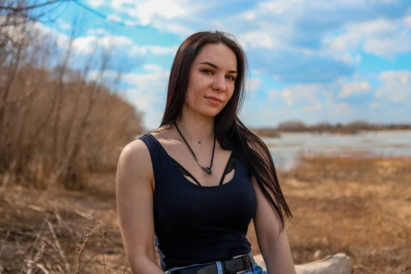 A beautiful girl is sitting on a log by the river Bank in jeans and a black t shirt