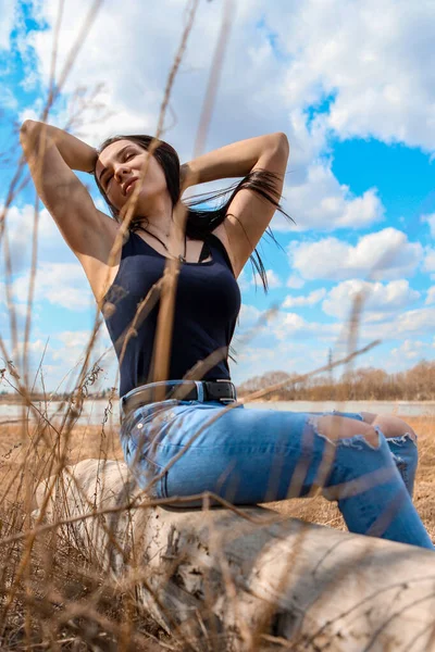 Güzel bir kız nehir kıyısında bir kütüğün üzerinde oturuyor kot pantolon ve siyah tişörtle.