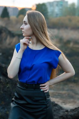 Mavi bluz ve deri etek giyen muhteşem bir bayan. Mavi gözleri nazikçe yana bakıyor. Uzun sarı saçlar rüzgarda dalgalanıyor. Çimenlerin ve evlerin arka planında.