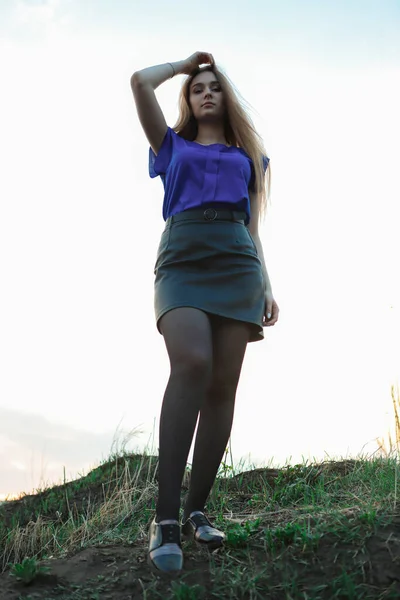 A tall, charming young girl with long blond hair, which she holds against the wind, stands on a hill with grass, against the light sky. Stylishly dressed in a blue blouse and black leather skirt.