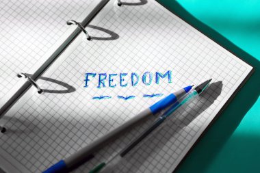 Kafesteki açık bir defterdeki yazıt özgürlüğü. Mavi ve yeşil kalemlerle yazılmış. Kalemler yazının yanındaki defterde..