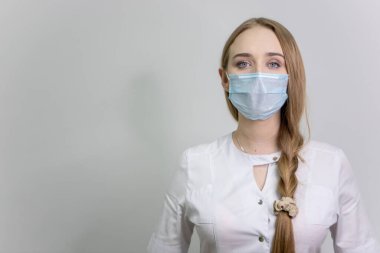 Tıbbi koruyucu maskeli ve beyaz önlüklü bir kız doktor kameraya bakar. Metin için boş alan. Beyaz bir duvara karşı. Saçları örgülü..