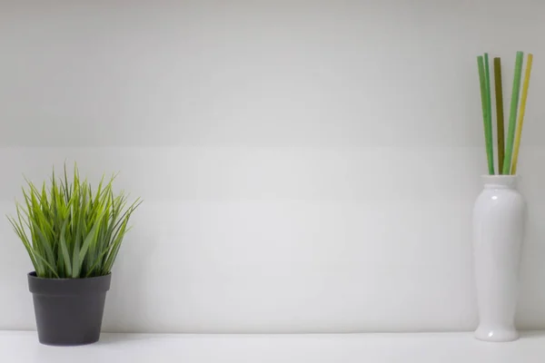Yeşil yapay çimenli siyah bir tencere ve bambu renkli çubuklu beyaz bir vazo. Beyaz bir duvara karşı beyaz bir rafta duruyorlar. Metin yazmak için boş bir alan var.