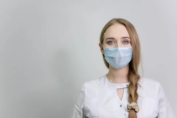 Tıbbi koruyucu maskeli ve beyaz önlüklü bir kız doktor kameraya bakar. Metin için boş alan. Beyaz bir duvara karşı. Saçları örgülü..