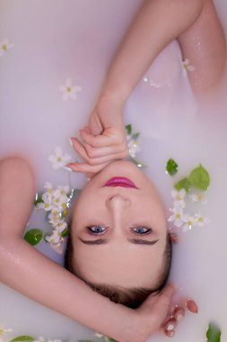 Zarif makyajlı ve güzel mavi gözlü çekici bir kız elma çiçekleri ve yapraklarıyla süslenmiş pembe bir renkle süt banyosu yapıyor. Nazik ellerle tatlı yüzüne dokunur.
