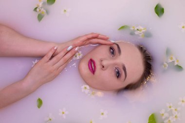 Zarif makyajlı ve güzel mavi gözlü çekici bir kız elma çiçekleri ve yapraklarıyla süslenmiş pembe bir renkle süt banyosu yapıyor. Nazik ellerle tatlı yüzüne dokunur.