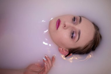 Zarif makyajlı ve güzel mavi gözlü çekici bir kız elma çiçekleri ve yapraklarıyla süslenmiş pembe bir renkle süt banyosu yapıyor. Nazik ellerle tatlı yüzüne dokunur.