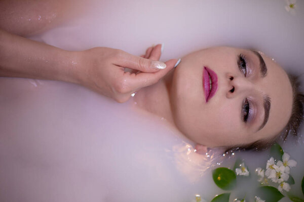 Очаровательная девушка со стильным макияжем и красивыми голубыми глазами принимает молочную ванну с розовым оттенком, усыпанным цветами и листьями яблок. Прикасается к сладкому лицу нежными руками
.