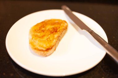 Beyaz ekmek hamurlu tost, tereyağı, gümüş bıçaklı beyaz tabak ve siyah granit tezgah.