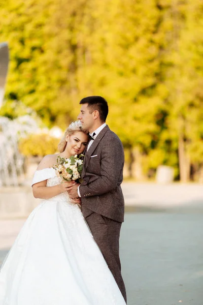 Bruden och brudgummen går tillsammans i parken. Charmig brud i en vit klänning, brudgummen är klädd i en mörk elegant kostym — Stockfoto