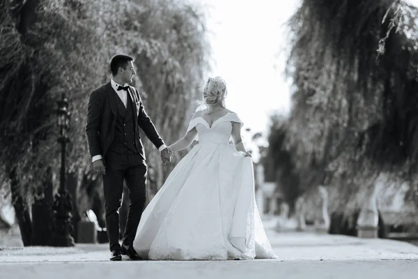 Les mariés marchent ensemble dans le parc. Mariée charmante dans une robe blanche, le marié est habillé d'un costume élégant sombre — Photo