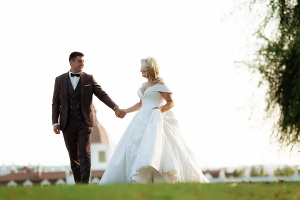 Les mariés marchent ensemble dans le parc. Mariée charmante dans une robe blanche, le marié est habillé d'un costume élégant sombre — Photo