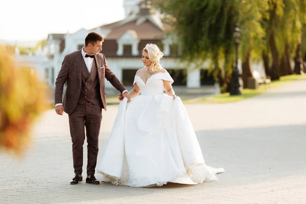 Gelin ve damat parkta birlikte yürürler. Beyaz elbiseli çekici gelin, damat koyu renkli şık bir takım giyiyor. — Stok fotoğraf