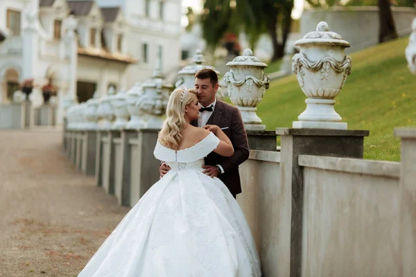 Η νύφη και ο γαμπρός περπατούν μαζί στο πάρκο. Γοητευτική νύφη με λευκό φόρεμα, ο γαμπρός είναι ντυμένος με ένα σκούρο κομψό κοστούμι — Φωτογραφία Αρχείου