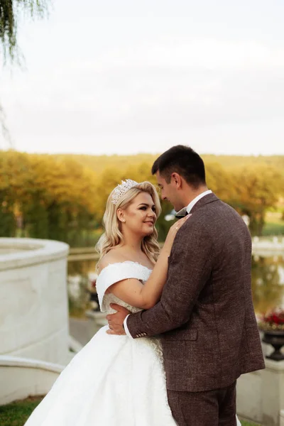 La novia y el novio caminan juntos en el parque. Encantadora novia en un vestido blanco, el novio está vestido con un traje elegante oscuro — Foto de Stock